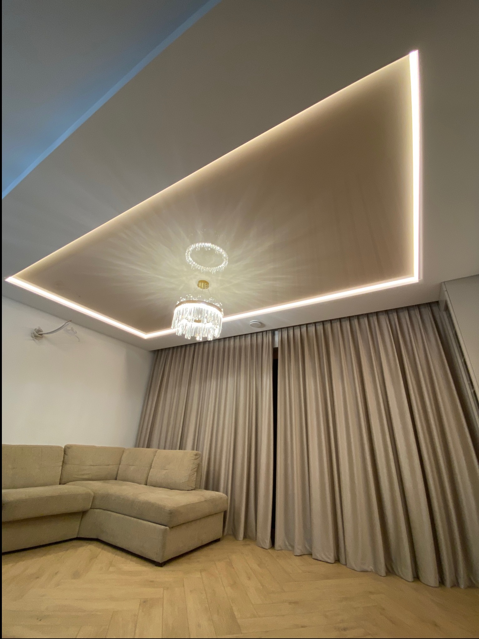 Salon z beżową, narożną sofą i podwieszanym sufitem z oświetleniem LED oraz kryształowym żyrandolem.