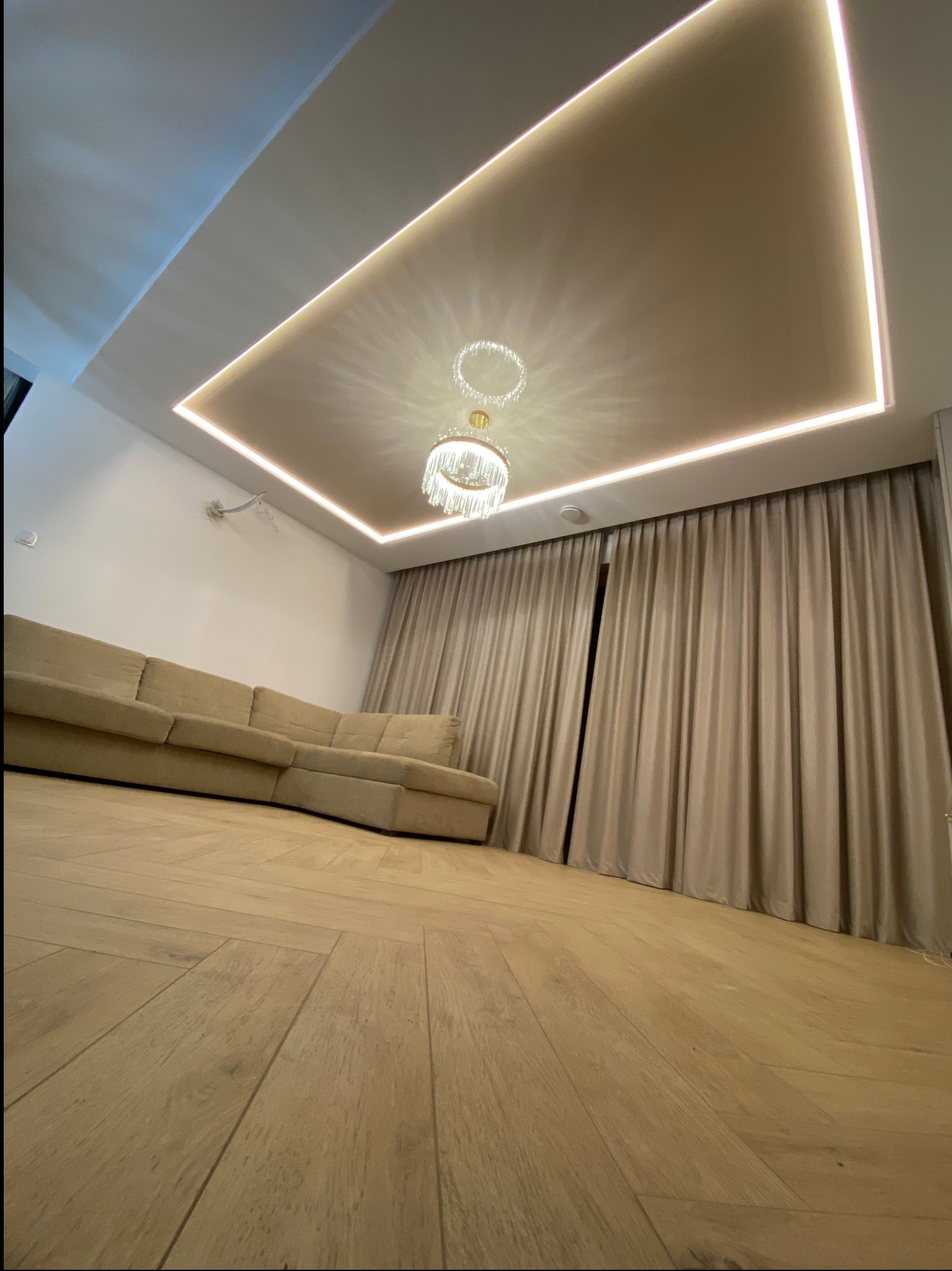 Salon z beżową sofą narożną, drewnianą podłogą w jodełkę i sufitem podwieszanym z oświetleniem LED oraz kryształowym żyrandolem, widok z dołu.
