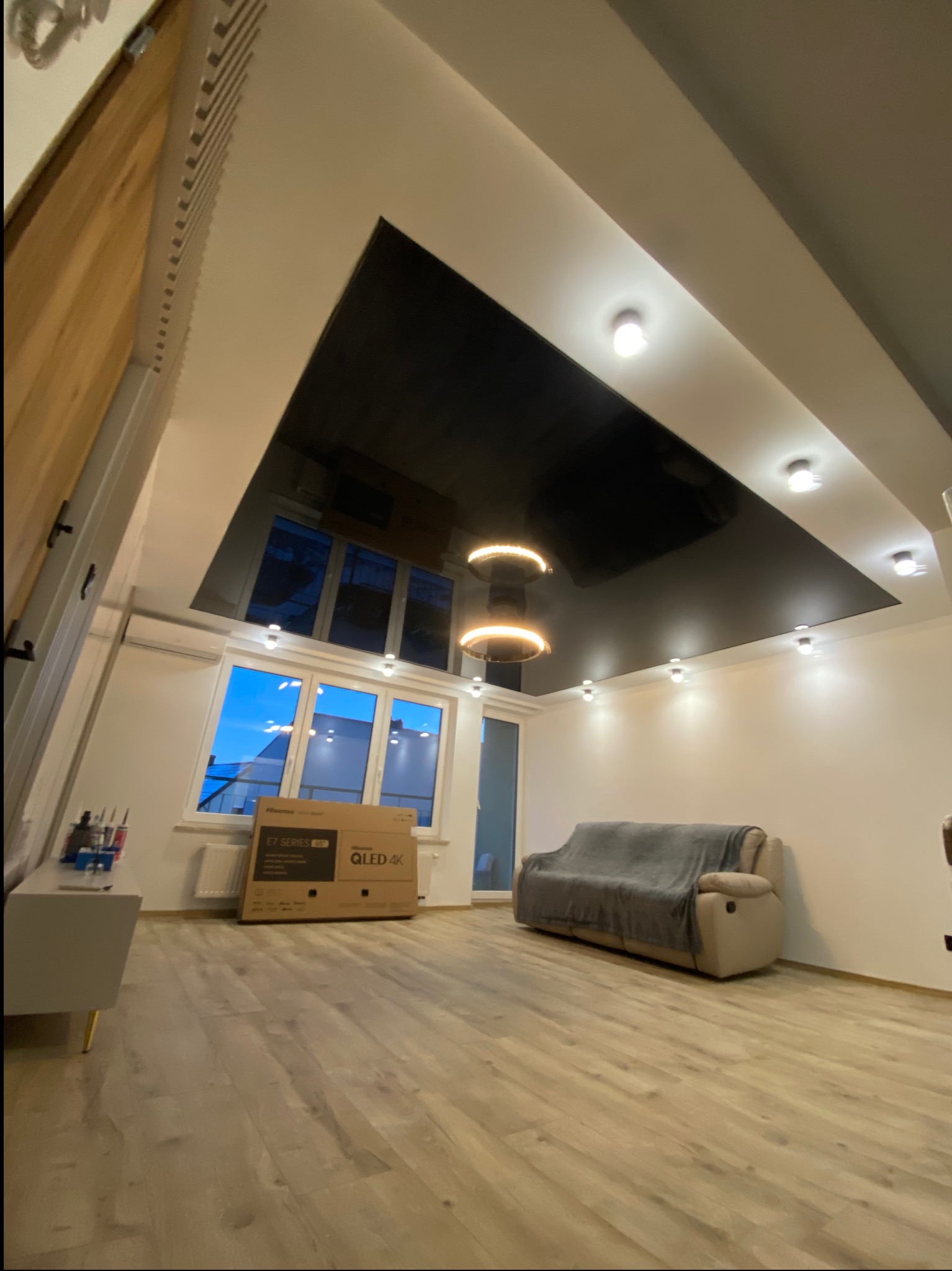 Salon z czarnym, błyszczącym sufitem napinanym, oświetleniem punktowym i nowoczesną lampą wiszącą, widoczne okno z widokiem na zewnątrz i podłoga z paneli.