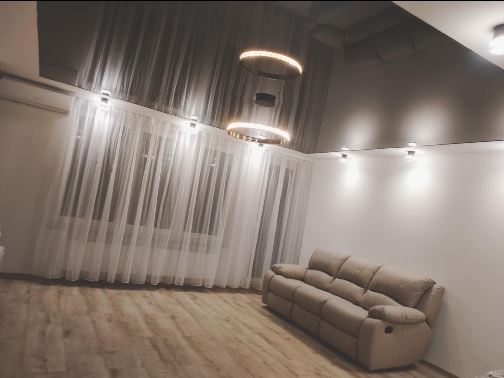 Salon z ciemnym, błyszczącym sufitem napinanym, okrągłymi lampami LED, jasnymi zasłonami i beżową sofą z funkcją relaks.