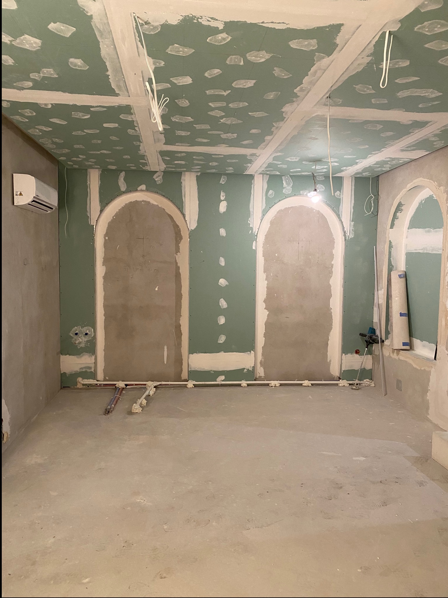 Pokój w trakcie prac wykończeniowych: ściany obłożone zielonymi płytami gipsowo-kartonowymi, widoczne szpachlowane łączenia, przygotowane łukowe wnęki, wystające przewody elektryczne i rury...