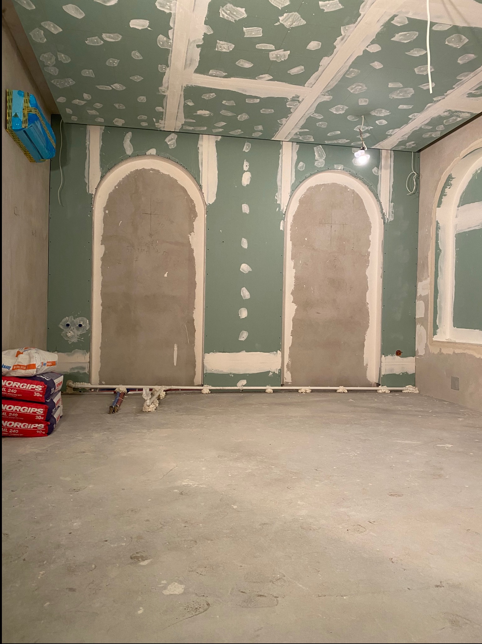 Wnętrze w trakcie remontu: ściany z płyt kartonowo-gipsowych, przygotowane do szpachlowania, z wykończonymi łukowatymi wnękami. Na podłodze worki z gipsem i rury grzewcze zabezpieczone pianką.