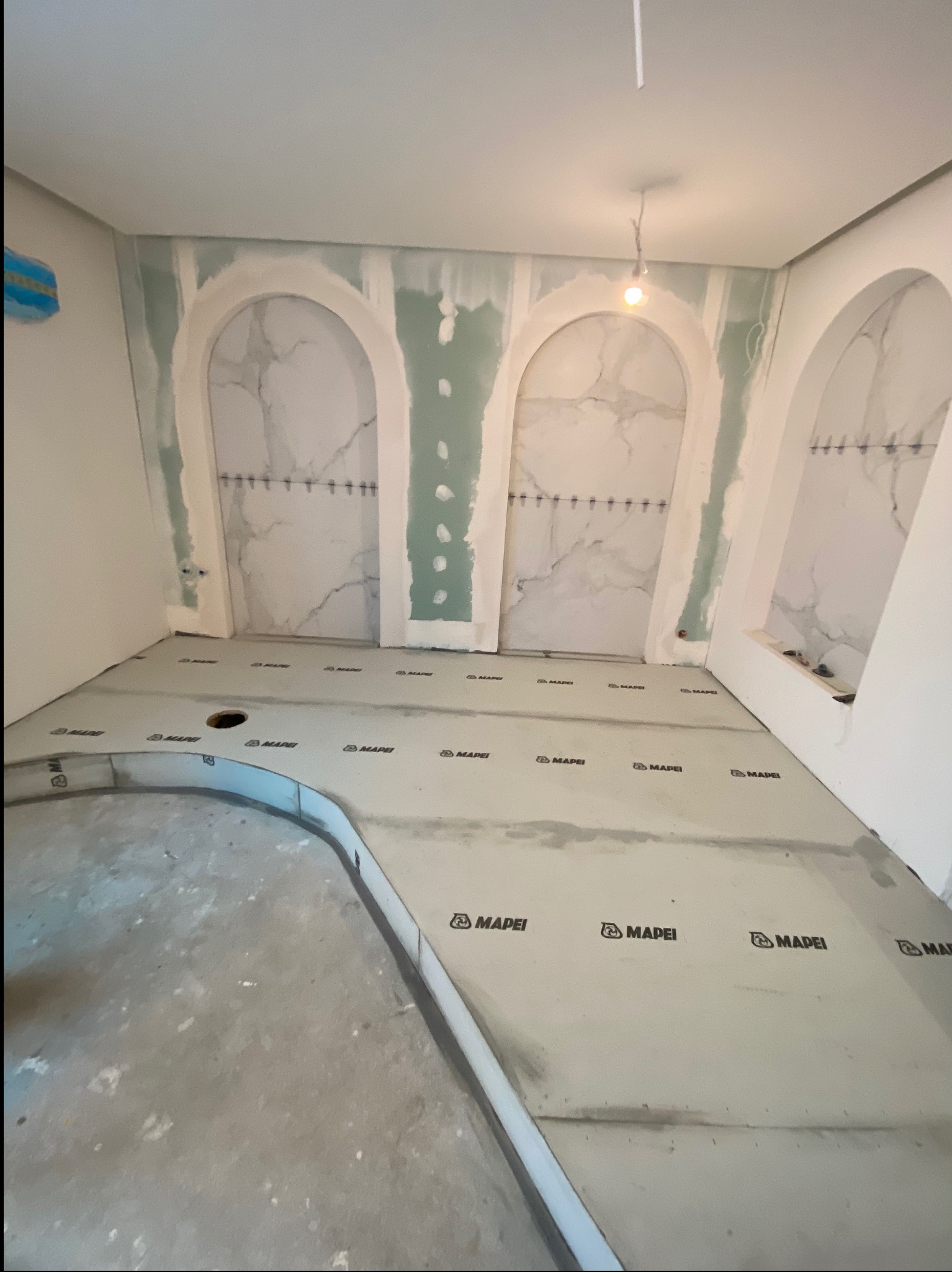 Łazienka w trakcie przygotowania do położenia płytek, widoczne wyprofilowane wnęki z imitacją marmuru, zabezpieczona podłoga z logo Mapei i odpływ liniowy.