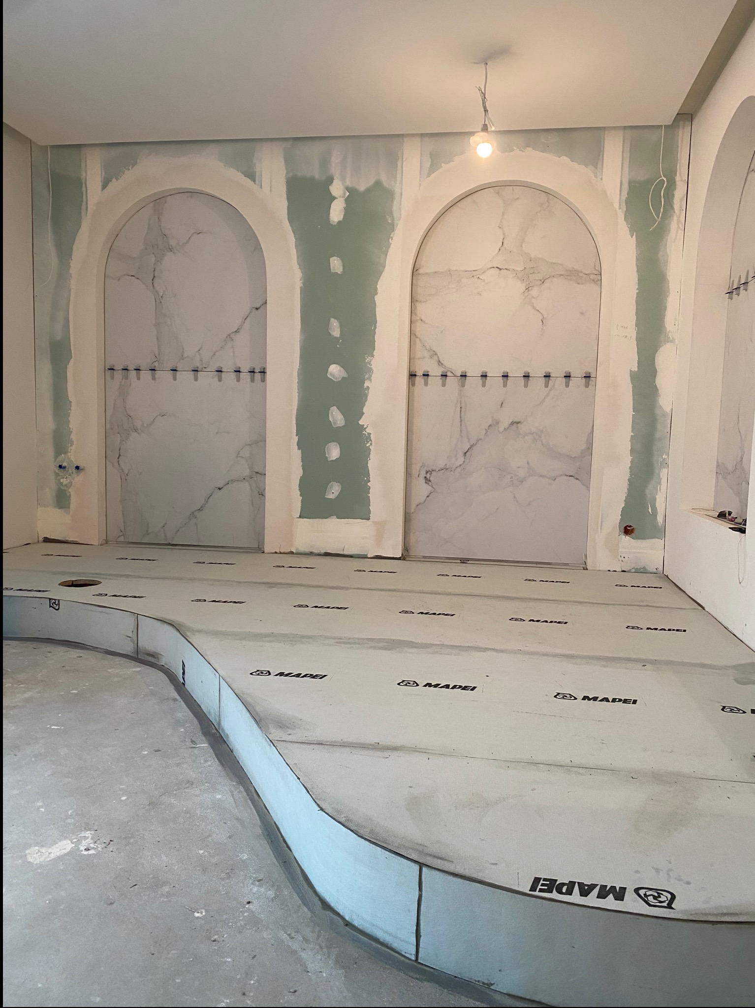 Wnętrze łazienki w trakcie prac wykończeniowych: widoczne przygotowane podłoże pod płytki z logo Mapei, wnęki w ścianach z imitacją marmuru, częściowo wykończone płytami gipsowo-kartonowymi.