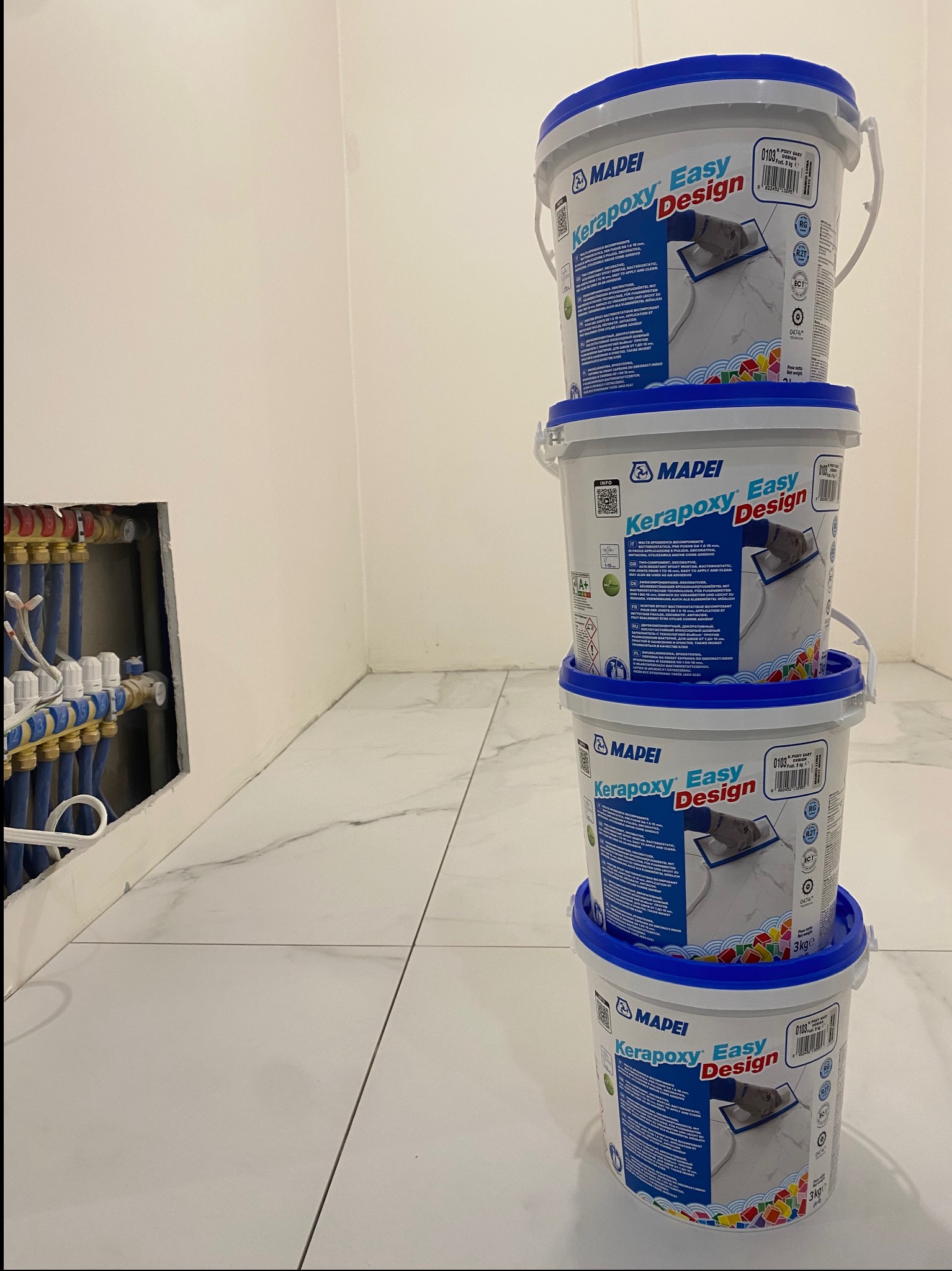 Stos czterech wiader z epoksydową zaprawą do spoinowania płytek, marki Mapei Kerapoxy Easy Design, na tle wykończonej białymi płytkami podłogi i fragmentu instalacji hydraulicznej w ścianie.