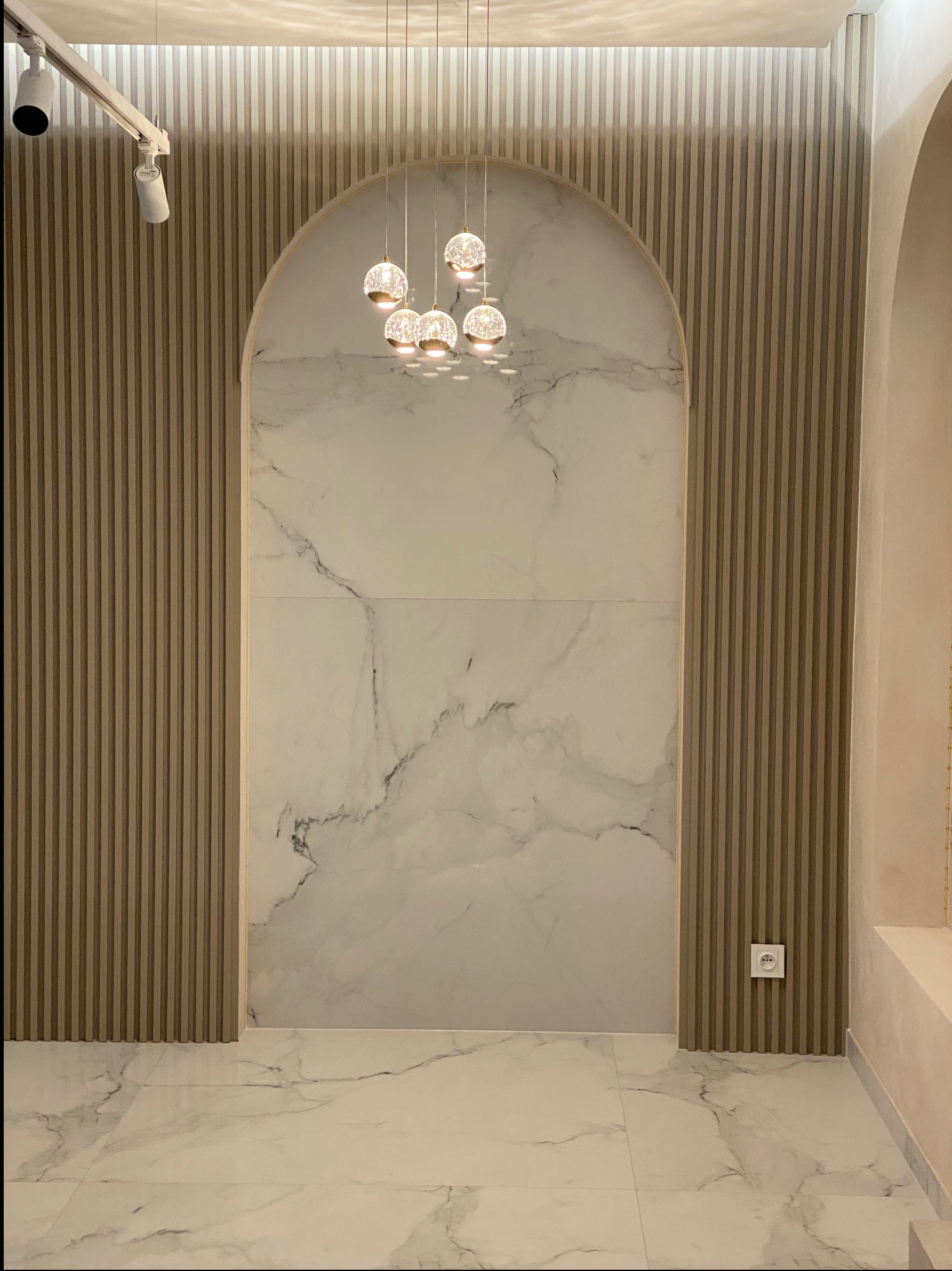 Pusta przestrzeń w pokoju z marmurową ścianą w kształcie łuku, oświetlona wiszącymi kulami, z dekoracyjnymi pionowymi panelami na ścianach bocznych i gładką, marmurową podłogą.