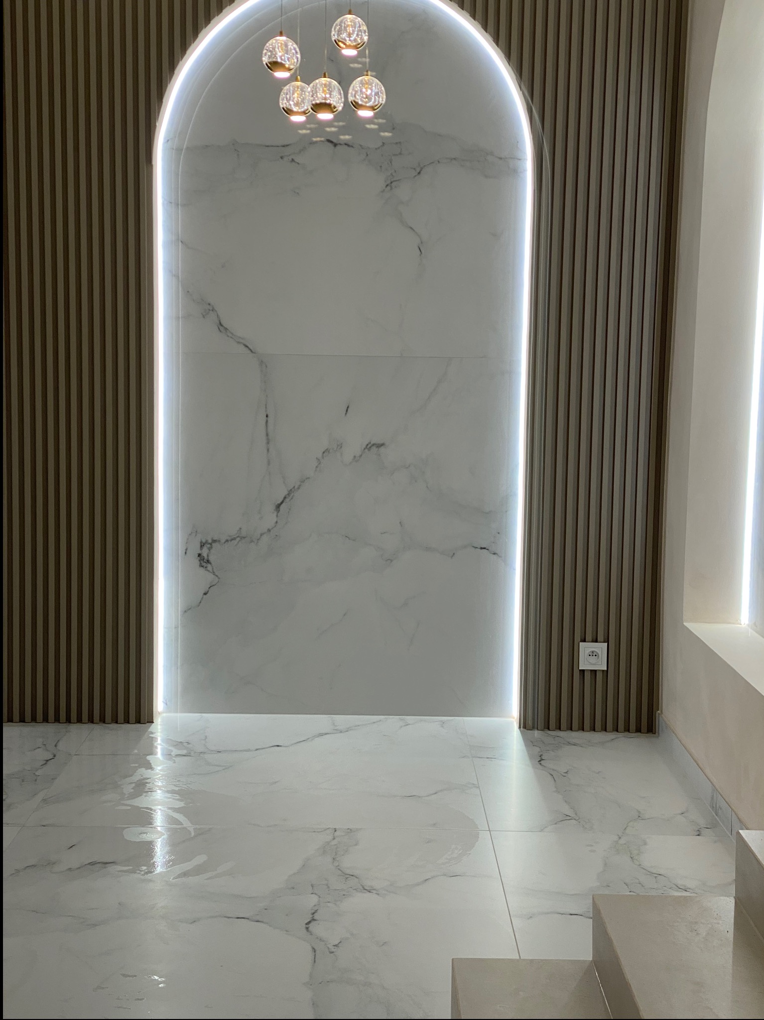 Nowoczesne wnętrze z marmurową ścianą w kształcie łuku, podświetloną listwą LED, drewnianymi panelami ściennymi i ozdobną lampą wiszącą z kilkoma szklanymi kloszami.