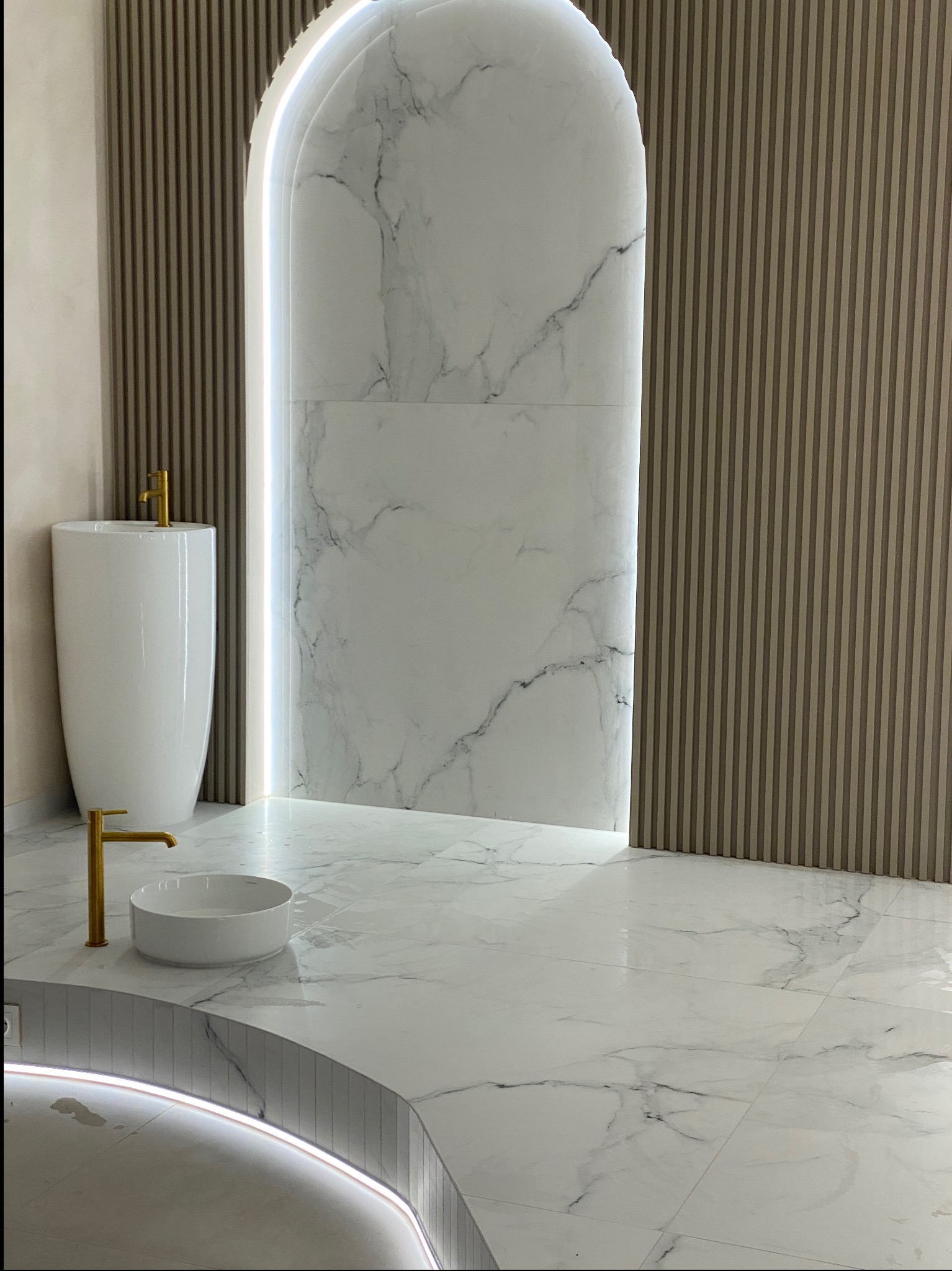 Nowoczesna łazienka z białymi płytkami imitującymi marmur, złotą armaturą, wolnostojącą umywalką i podświetlanym łukiem w ścianie z drewnianymi panelami.