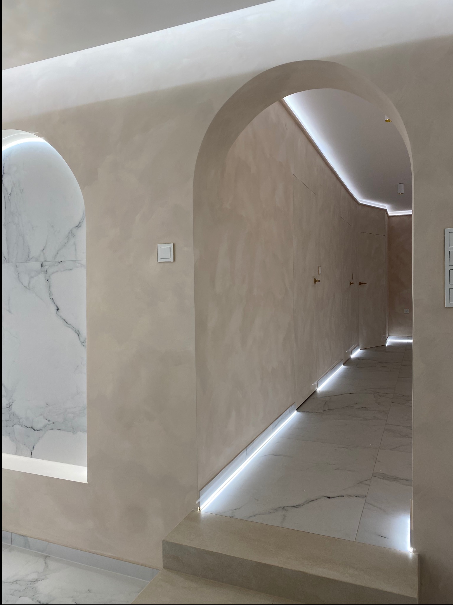 Elegancki korytarz z łukowymi przejściami, kremowymi ścianami z fakturą, marmurową wnęką, podświetleniem LED przy podłodze i suficie, oraz marmurową podłogą.