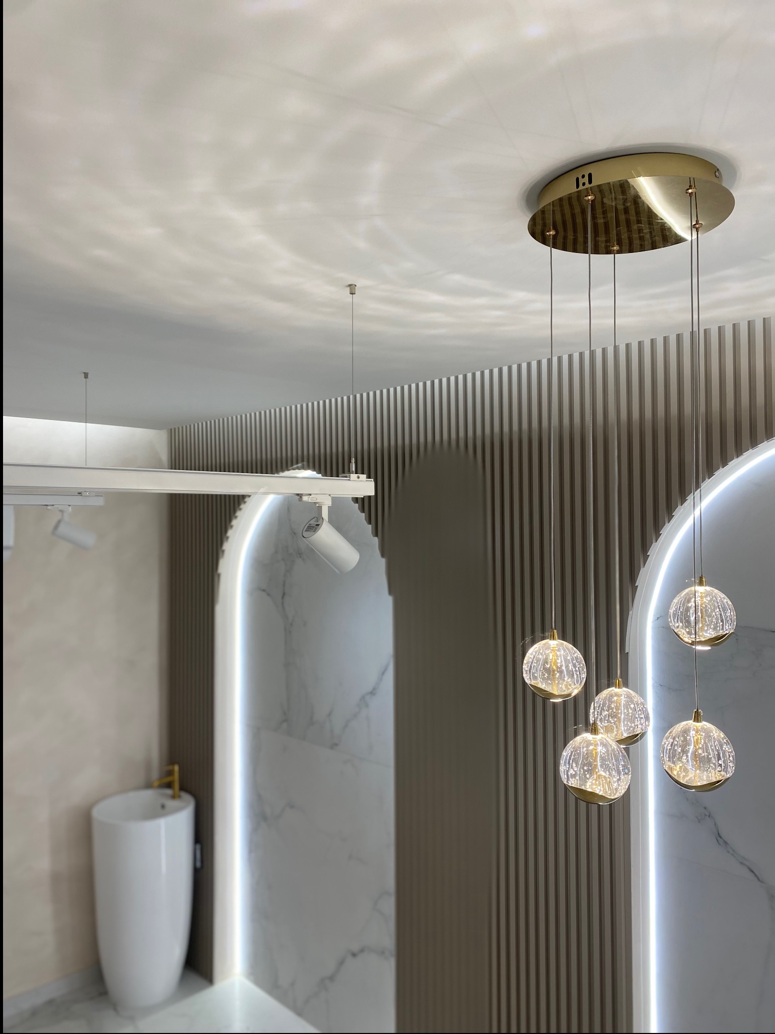 Nowoczesna łazienka z podświetlanym lustrem w kształcie łuku, wiszącą lampą z kulistymi kloszami i wolnostojącą umywalką z baterią w kolorze złota.