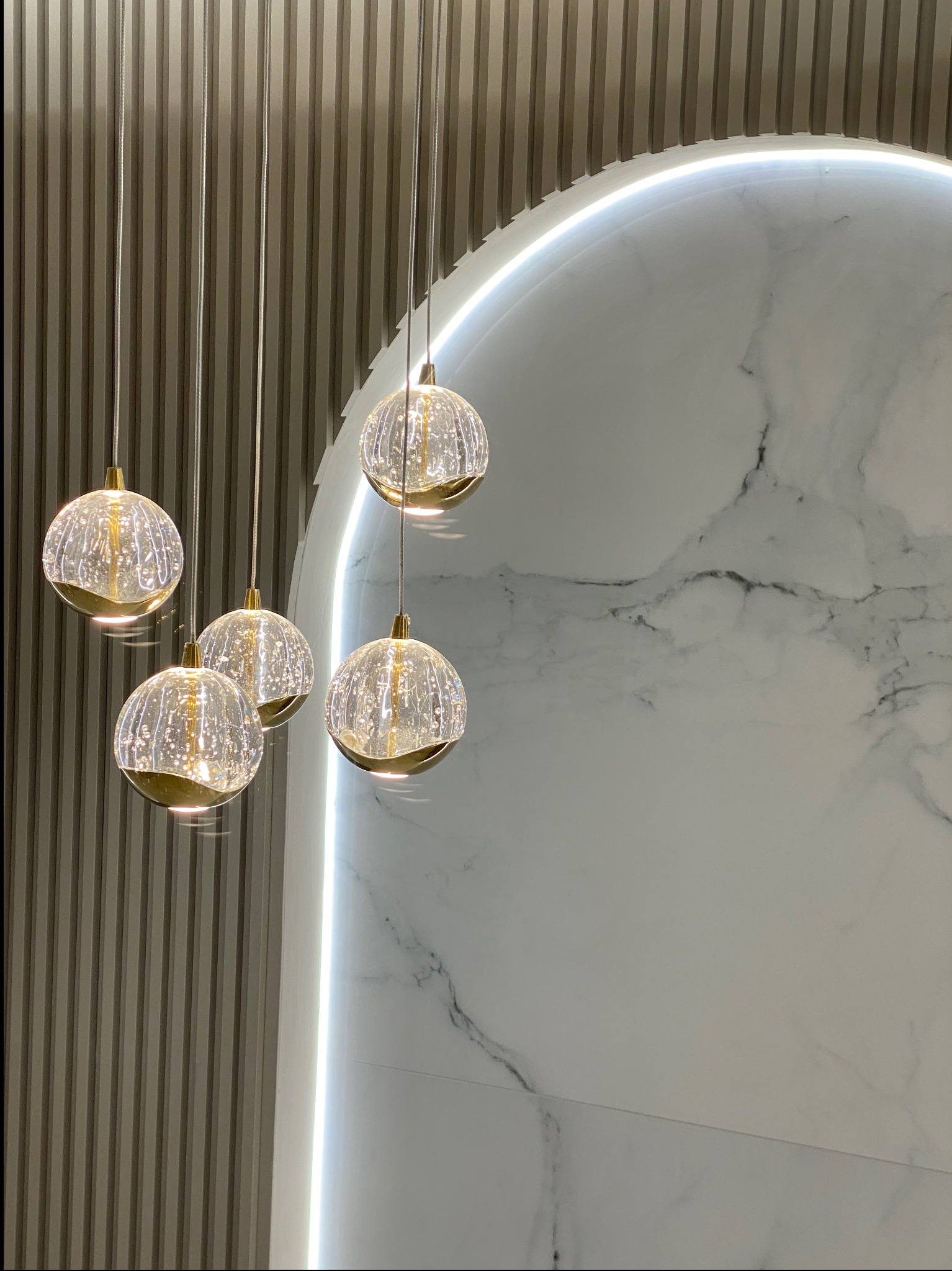 Cztery szklane lampy wiszące o kulistym kształcie z złotymi akcentami, na tle ściany z pionowymi panelami i marmurowej płyty z podświetleniem LED w kształcie łuku.