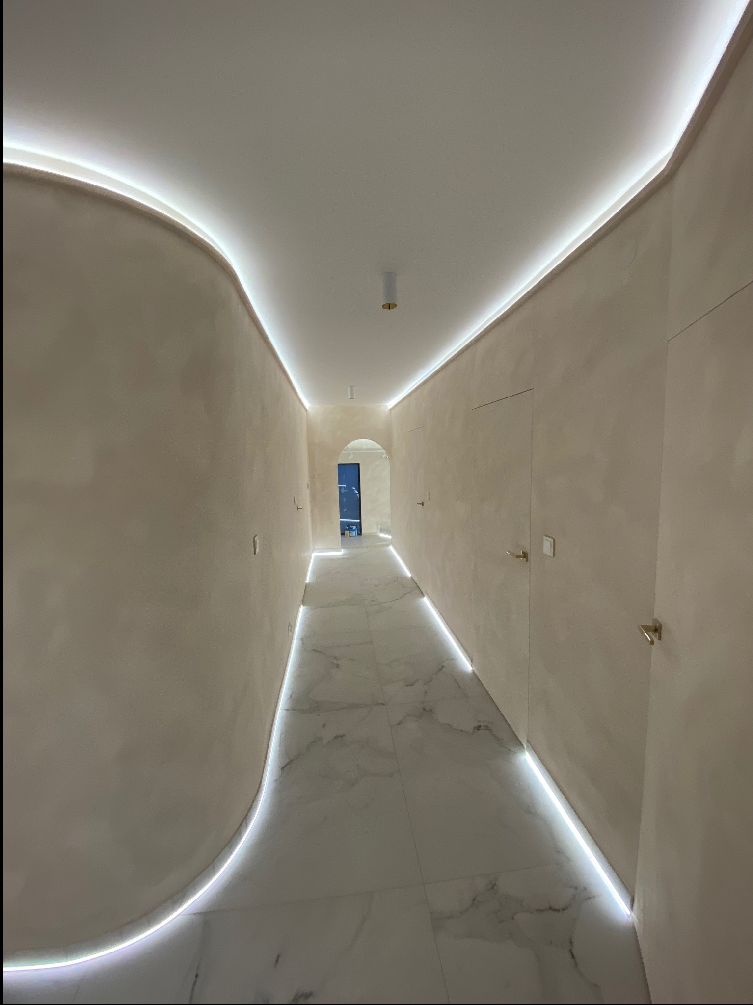 Korytarz z beżowymi ścianami o nieregularnej fakturze, białymi marmurowymi płytkami na podłodze i oświetleniem LED wzdłuż sufitu i podłogi, tworzącym nowoczesny efekt.