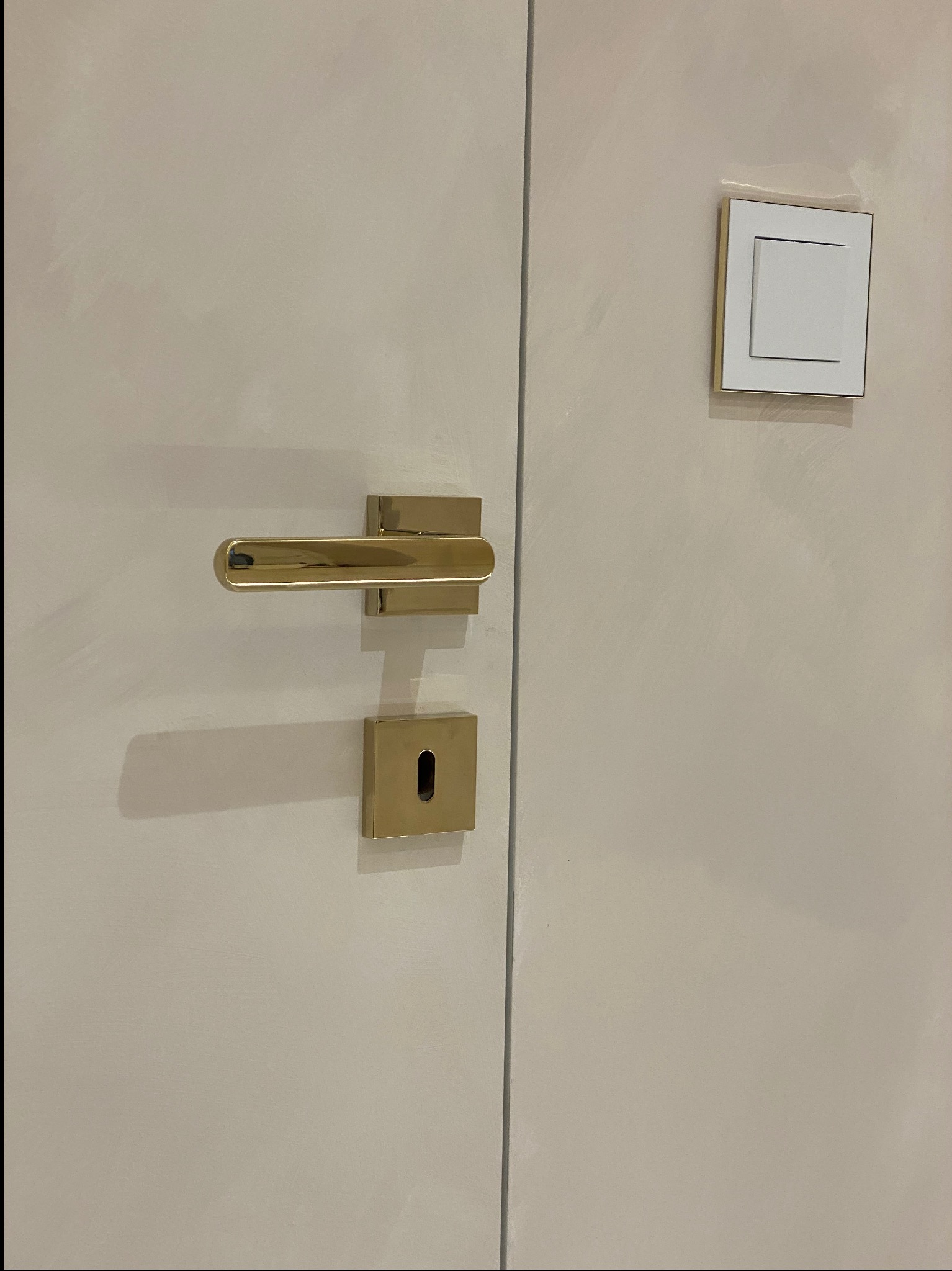 Detal zdjęcia: kremowe drzwi z złotą klamką i zamkiem, obok biały włącznik światła z złotą ramką, widoczne ślady malowania na ścianie.