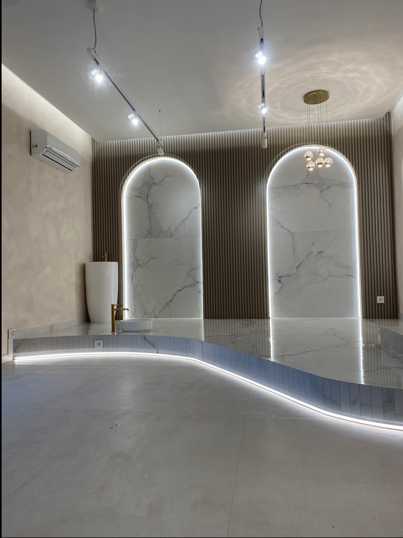 Nowoczesna łazienka z podwyższoną podłogą wykończoną płytkami imitującymi marmur, oświetleniem LED wzdłuż krawędzi, umywalką wolnostojącą i dwoma wnękami w ścianie z dekoracyjnymi panelami.