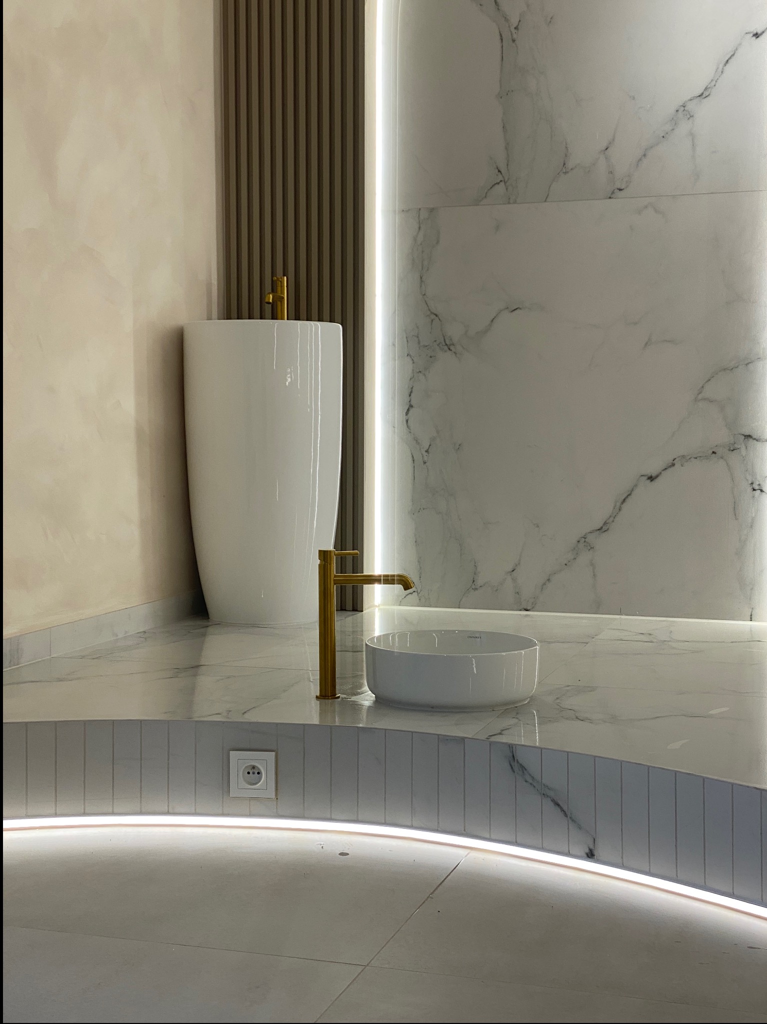 Nowoczesna łazienka z wolnostojącą umywalką, złotą armaturą i marmurowymi płytkami, podświetlana listwa LED w dolnej części ściany, gładkie ściany w beżowym odcieniu.