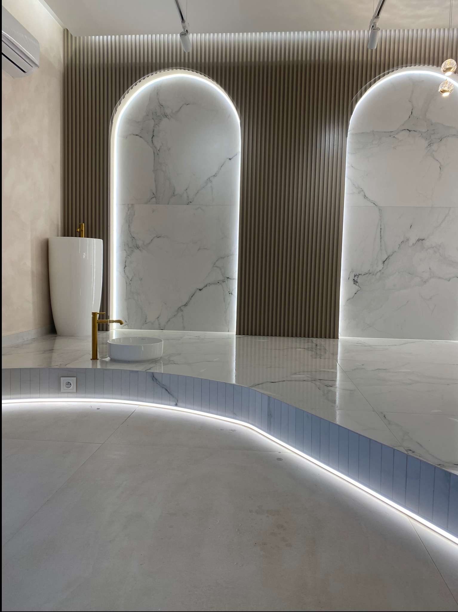 Nowoczesna łazienka z podświetlanym podestem, marmurowymi płytami w kształcie łuków i wolnostojącą umywalką w kolorze białym z złotą armaturą.