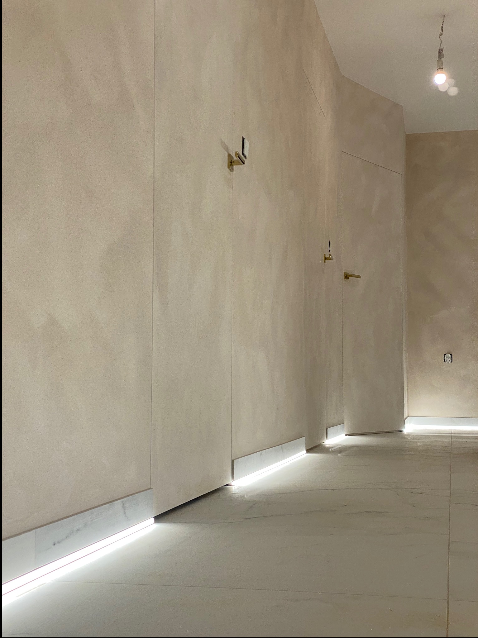 Beżowe ściany z dekoracyjnym tynkiem i ukrytymi drzwiami z złotymi klamkami, podświetlenie LED przy podłodze, marmurowe płytki podłogowe.