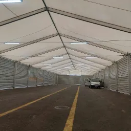 Wnętrze dużego, pustego namiotu magazynowego z oświetleniem LED pod sufitem, szarymi ścianami z blachy falistej i asfaltową podłogą z żółtymi liniami; otwarte drzwi samochodu dostawczego po prawej...