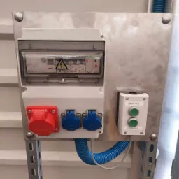 Srebrna płyta montażowa z zabezpieczeniami elektrycznymi, gniazdami przemysłowymi (czerwone i dwa niebieskie) oraz dwoma przyciskami (Sekcja 1 i Sekcja 2) w białej obudowie. Widoczne niebieskie...