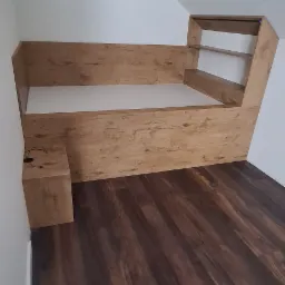 Drewniana zabudowa łóżka z półkami w skosie poddasza, dopasowana do ciemnej podłogi, z małą szafką nocną po lewej stronie.