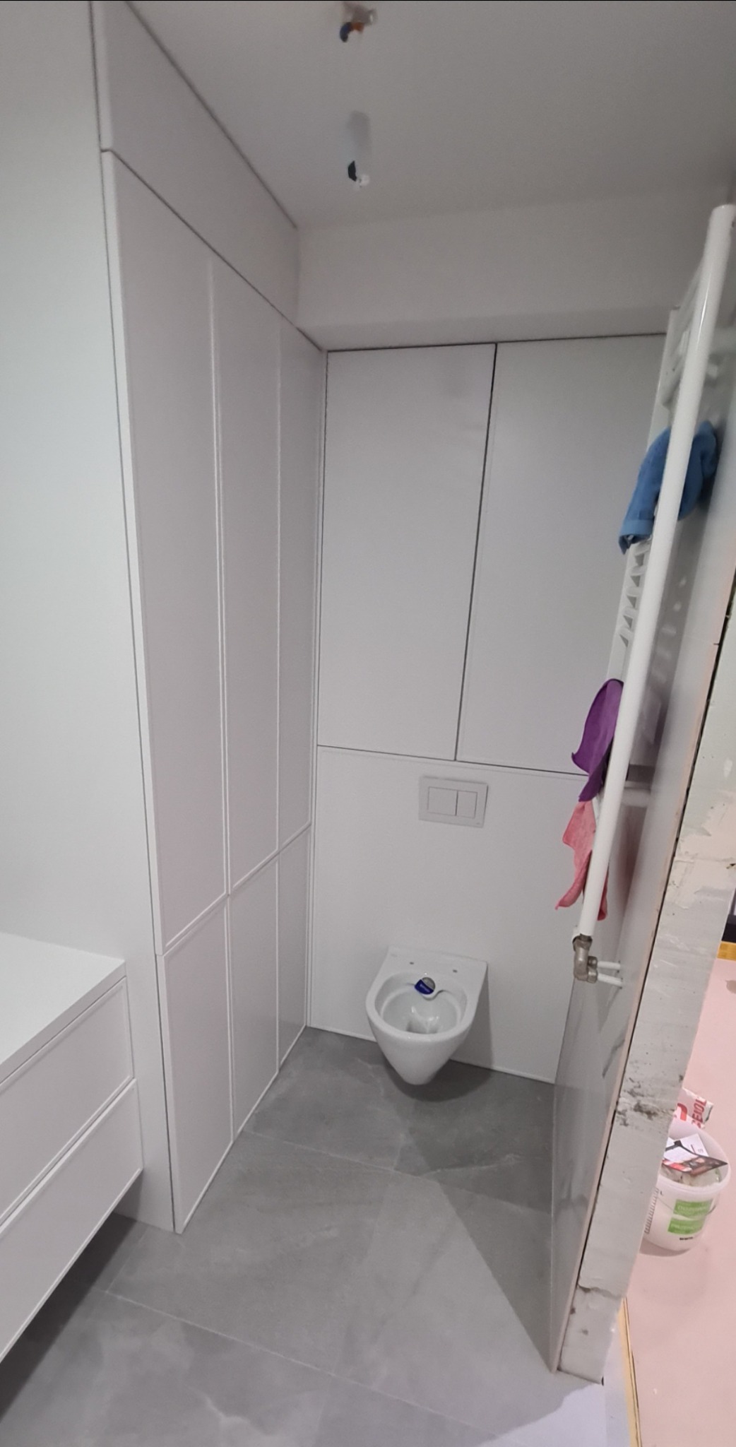 Biała zabudowa w małej łazience z szarą podłogą, widoczna toaleta i grzejnik z powieszonymi ręcznikami, częściowo widoczne akcesoria malarskie.