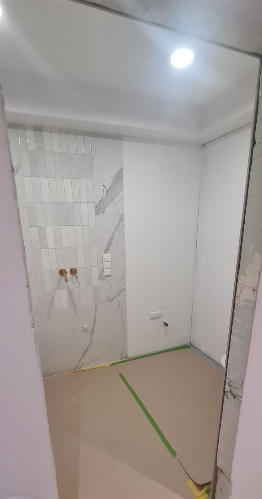 Wnętrze łazienki w trakcie remontu, widoczne płytki imitujące marmur na ścianie, złote baterie umywalkowe, podłoga zabezpieczona taśmą malarską, surowy stan wykończenia.