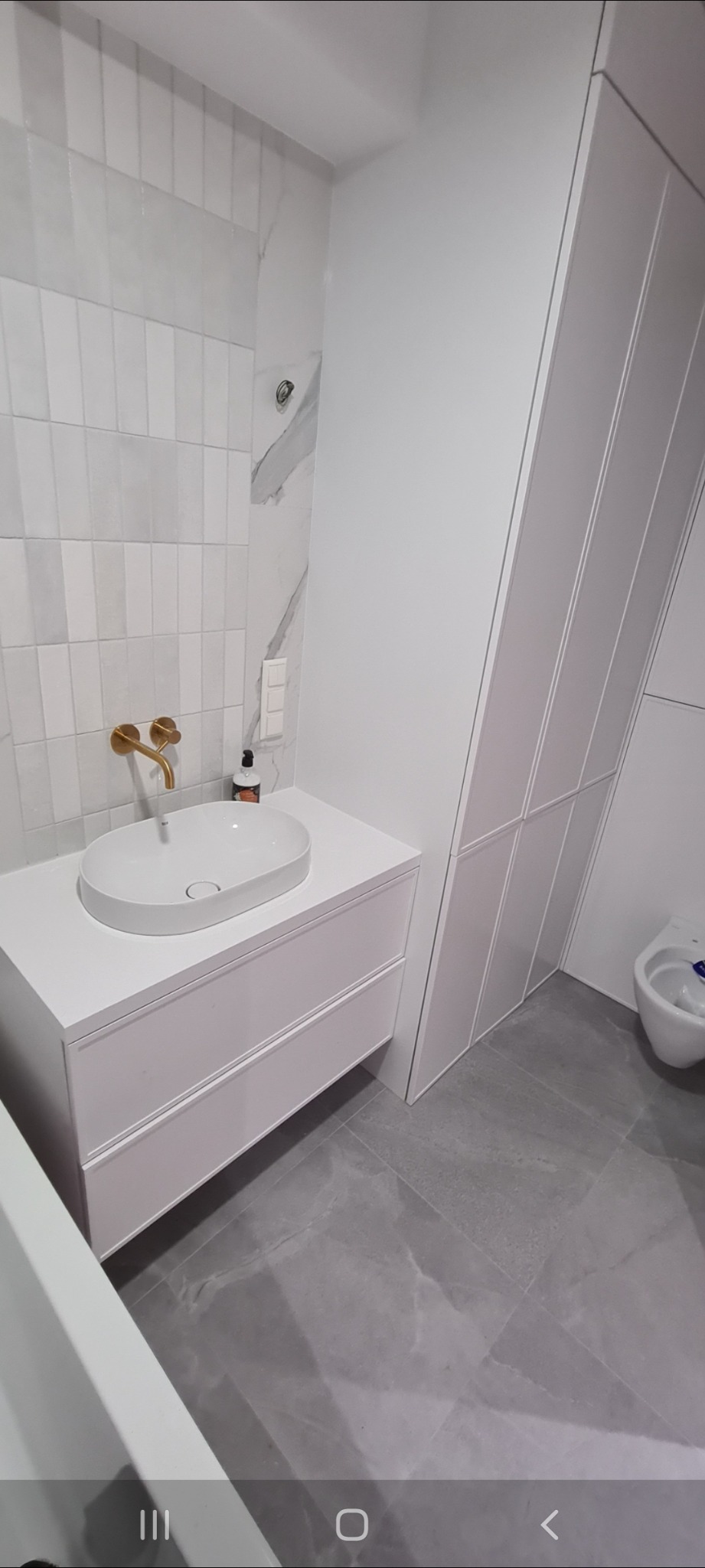 Nowoczesna łazienka z białą umywalką nablatową, złotą armaturą i szafką pod umywalką w zabudowie, uzupełniona płytkami imitującymi marmur i wysoką szafą.