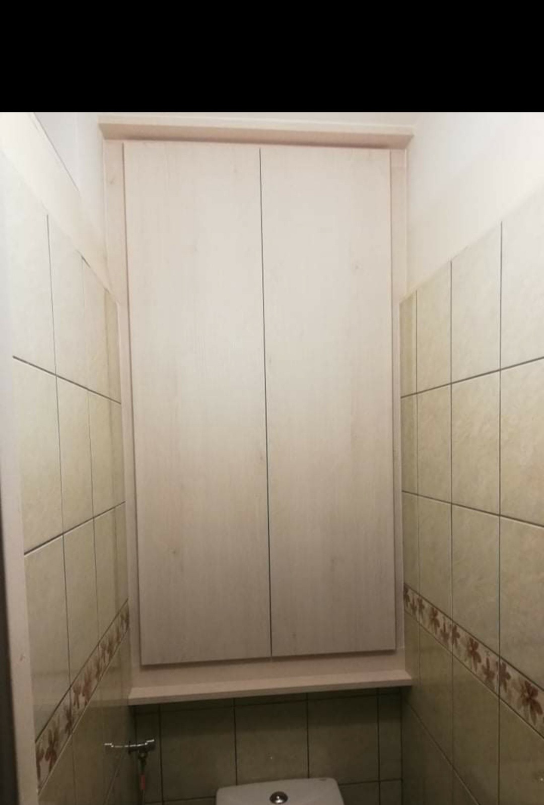 Biała szafka z dwoma drzwiczkami zamontowana nad toaletą w łazience wyłożonej jasnymi, kwadratowymi płytkami z dekoracyjnym paskiem.