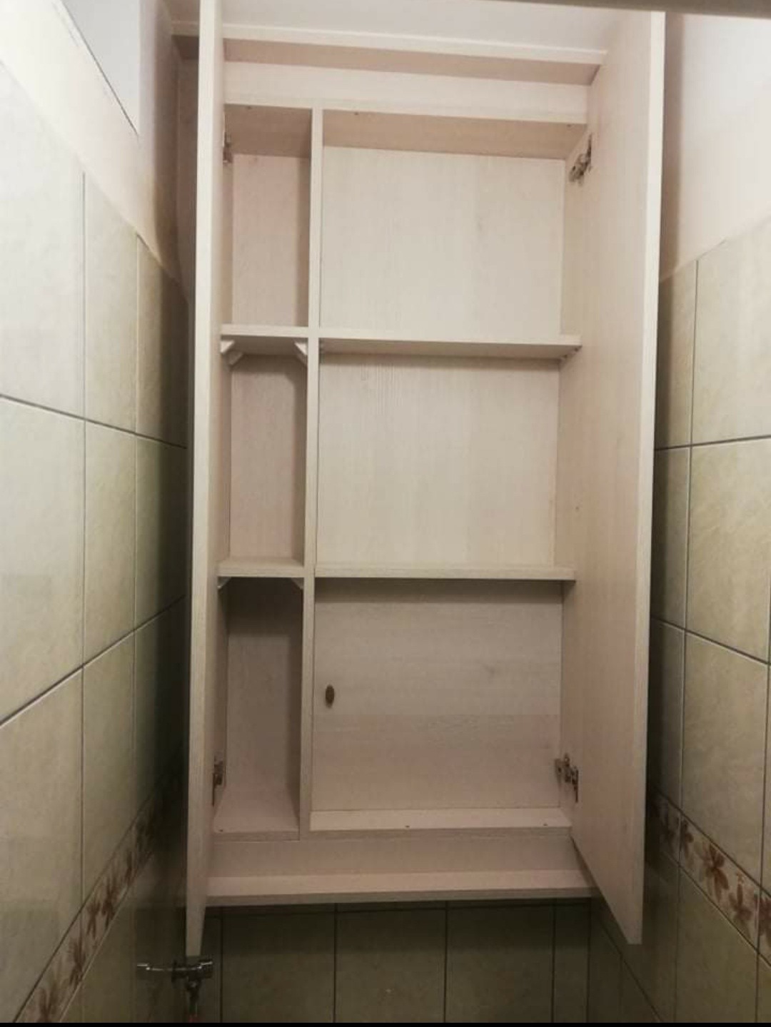 Otwarta, jasna szafka łazienkowa z półkami, zamontowana na kafelkowanej ścianie, widoczne zawiasy i uchwyt.