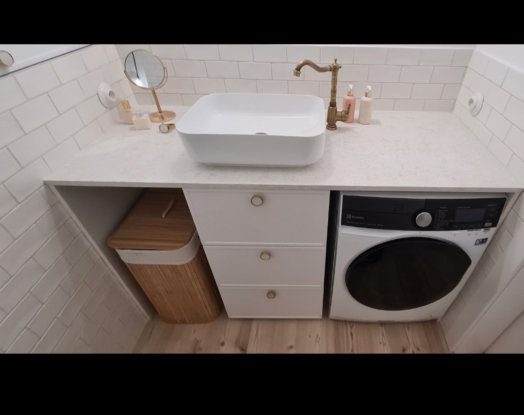 Nowoczesna łazienka z białą, prostokątną umywalką nablatową, baterią w kolorze starego złota, szafką z trzema szufladami, koszem na pranie i pralką zabudowaną pod blatem.