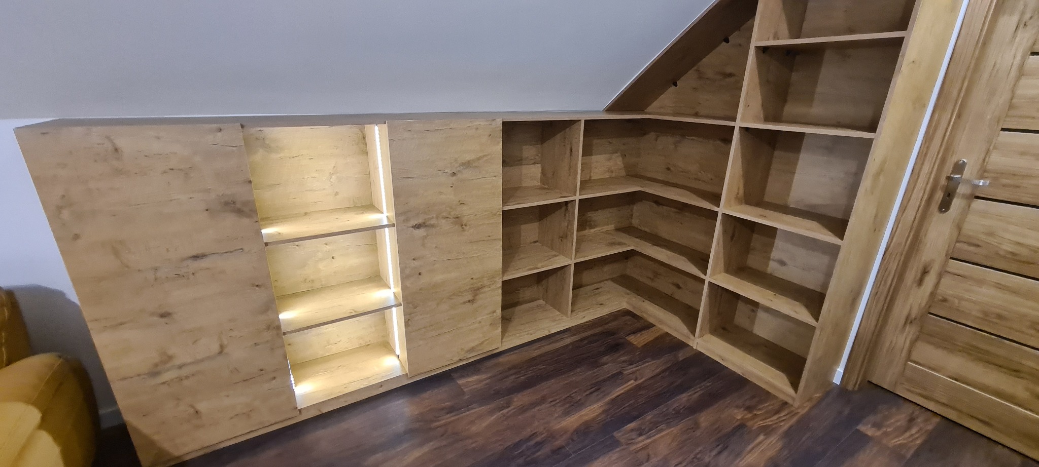 Szafa wnękowa na poddaszu z oświetleniem LED wewnątrz, wykonana z płyty drewnopodobnej, dopasowana do skosu dachu i drewnianej podłogi.