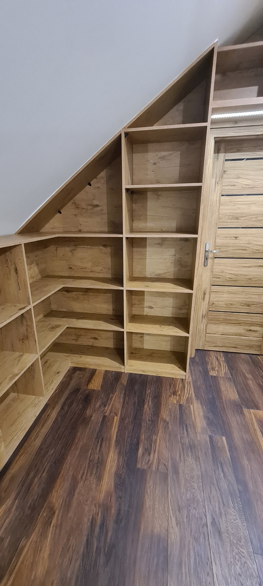 Drewniana zabudowa pod skosem z półkami i drzwiami, dopasowana do poddasza, z oświetleniem LED nad drzwiami, na podłodze panele w odcieniach brązu.