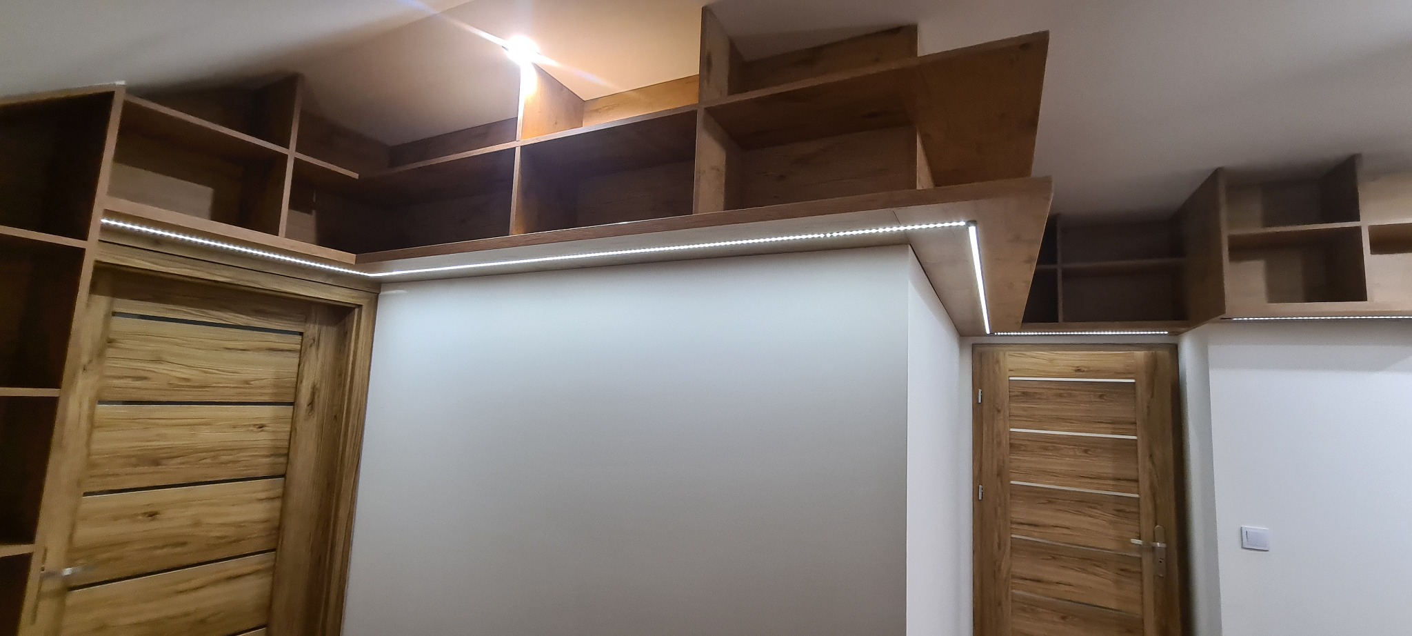 Drewniane półki nad drzwiami z oświetleniem LED, tworzące zabudowę wnęki, widok od dołu.