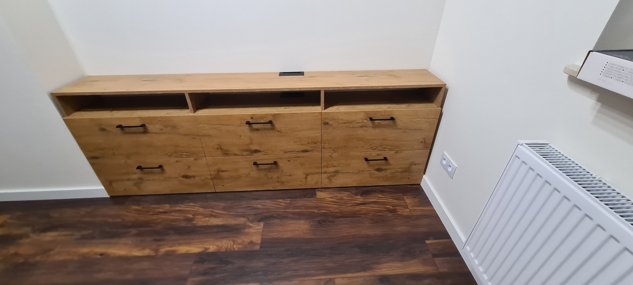 Drewniana szafka RTV z trzema szufladami i otwartymi półkami, z czarnymi uchwytami, umieszczona na brązowej podłodze w pomieszczeniu z białymi ścianami i grzejnikiem.