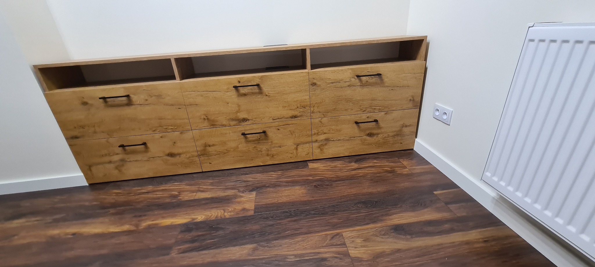 Drewniana komoda z szufladami i otwartymi półkami, widoczne czarne uchwyty, brązowa podłoga imitująca drewno, biała ściana z gniazdkiem elektrycznym i grzejnikiem.