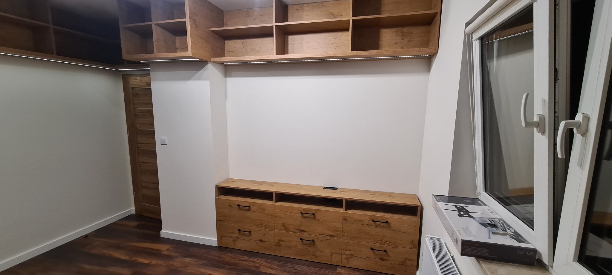 Drewniana szafa wnękowa z półkami i szafkami, oświetlenie LED pod półkami, widoczna drewniana podłoga, okno z roletą na parapecie leży pudełko z uchwytem do telewizora.