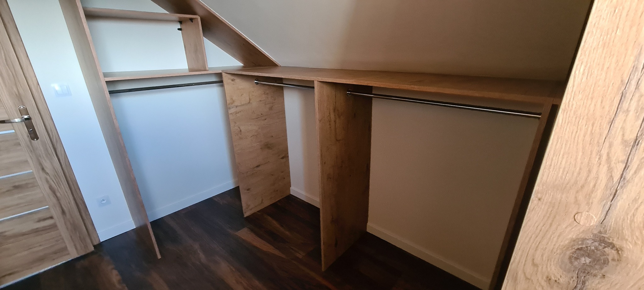 Zabudowa garderoby na poddaszu z drewnianymi elementami, widoczne drążki na ubrania i skośny sufit.