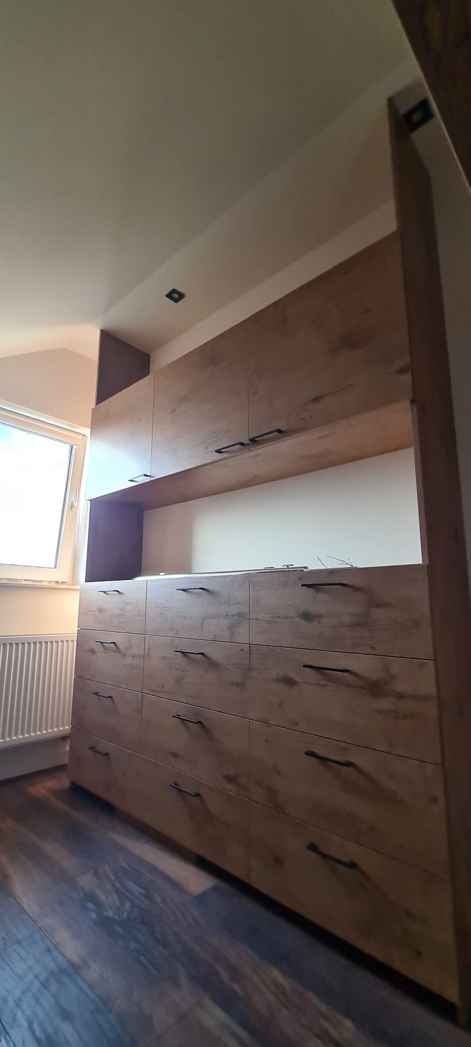 Drewniana szafa na wymiar z szufladami i górnymi szafkami, widoczna z perspektywy podłogi, z czarnymi uchwytami i oświetleniem punktowym na suficie.