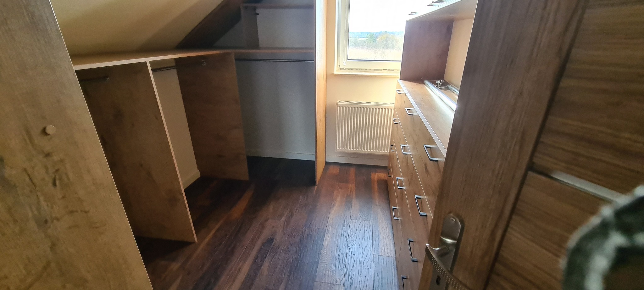 Korytarz garderoby na poddaszu z drewnianymi półkami, drążkiem na ubrania i komodą z szufladami, widok z perspektywy wejścia z widoczną klamką.