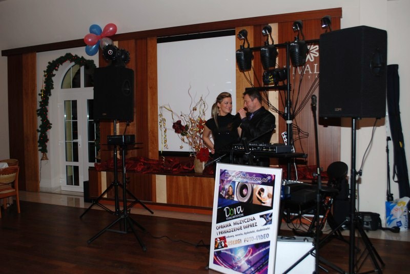 Wodzirej z mikrofonem i kobieta obok instrumentów muzycznych, dekoracja sali z oświetleniem i nagłośnieniem, oraz tablica reklamowa studia oprawy muzycznej.