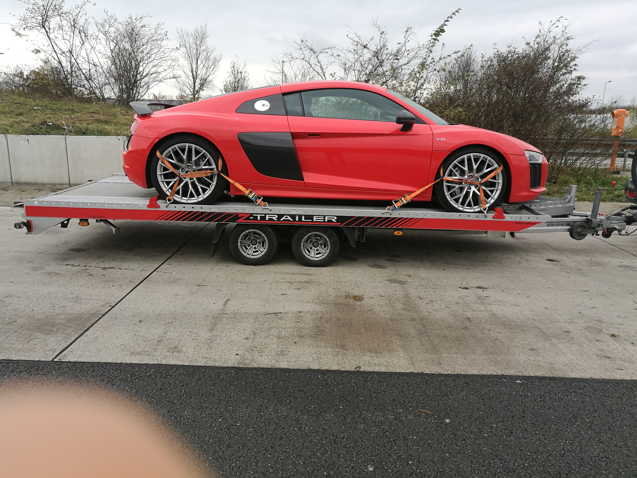 Czerwone sportowe Audi R8 V10 zabezpieczone pomarańczowymi pasami na srebrnej platformie lawety, widok z boku na tle szarego nieba i drzew bez liści.