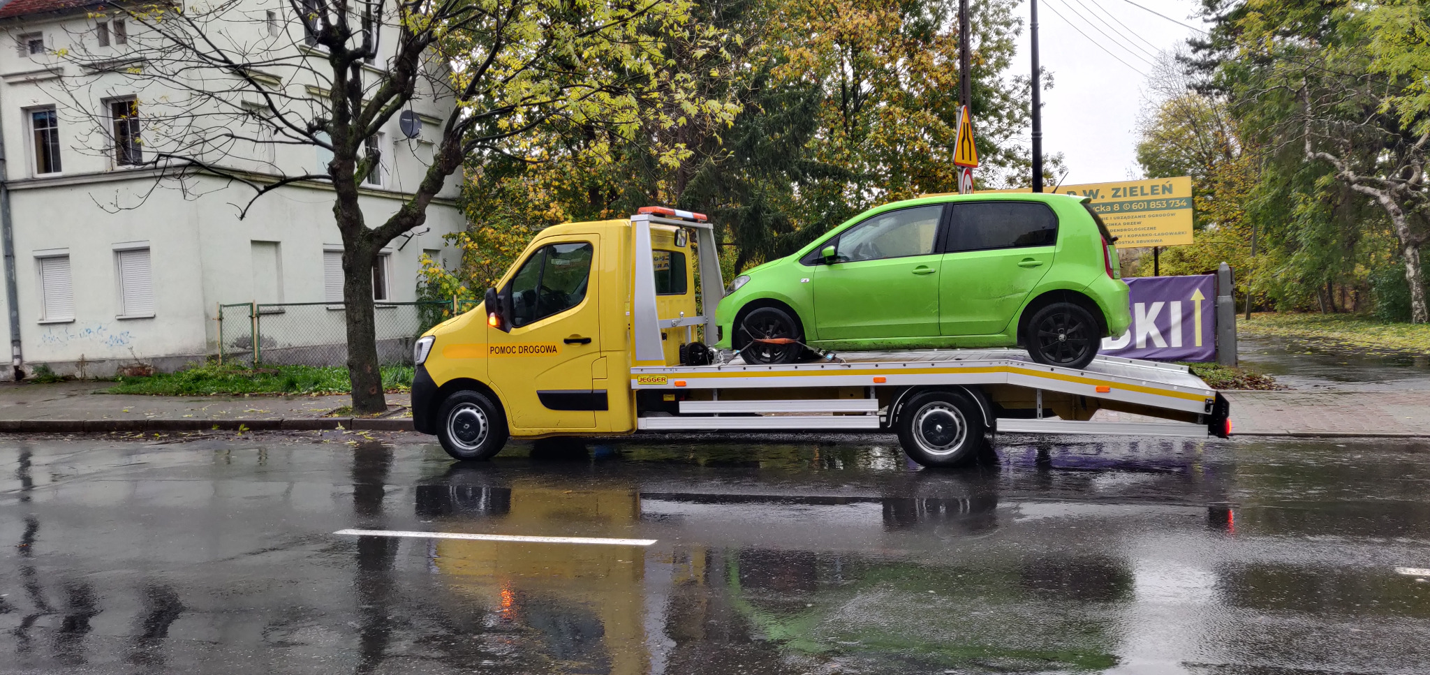 Żółta laweta z zielonym samochodem na platformie, zaparkowana na mokrej ulicy w jesienny dzień, widoczny budynek i drzewa w tle.