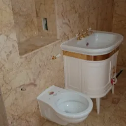 Wykończona łazienka z marmurowymi ścianami i podłogą, prezentująca białą toaletę z delikatnym kwiatowym wzorem na pokrywie oraz umywalkę w stylu retro z szafką i złotymi detalami.