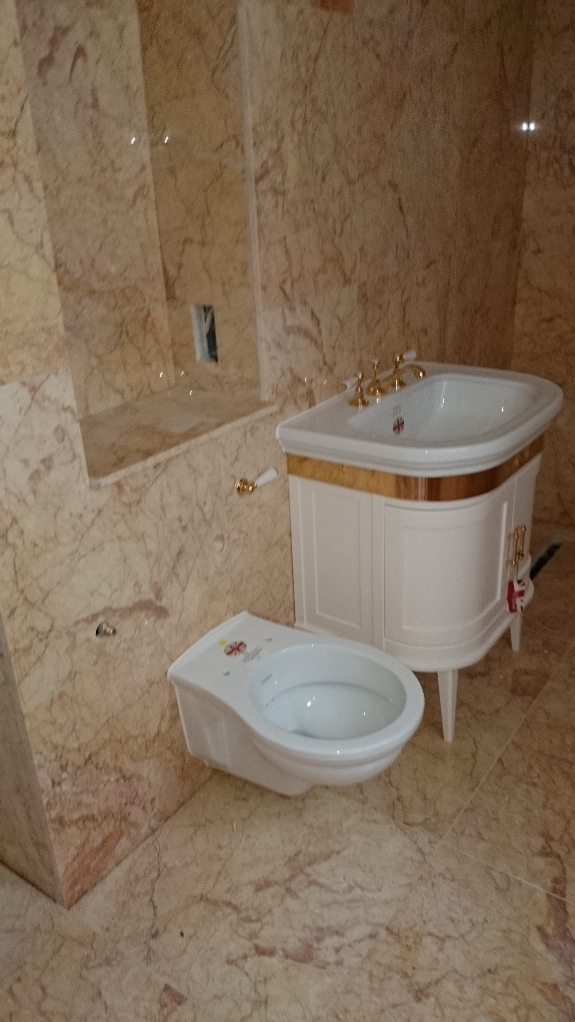 Wykończona łazienka z marmurowymi ścianami i podłogą, prezentująca białą toaletę z delikatnym kwiatowym wzorem na pokrywie oraz umywalkę w stylu retro z szafką i złotymi detalami.