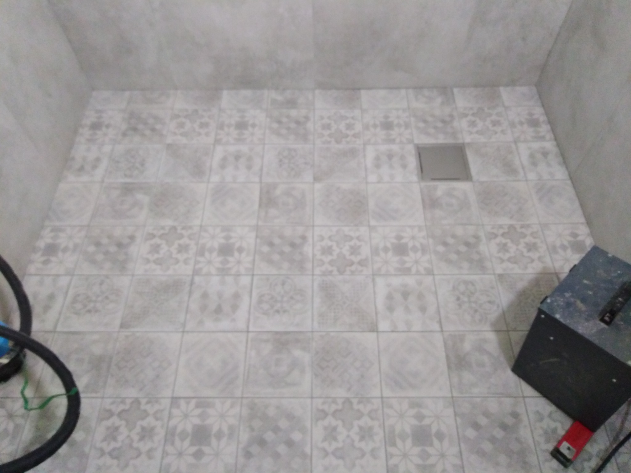 Świeżo ułożona, wzorzysta, szara podłoga z płytek w narożnej części łazienki, z widocznym odpływem liniowym i sprzętem budowlanym w rogu.