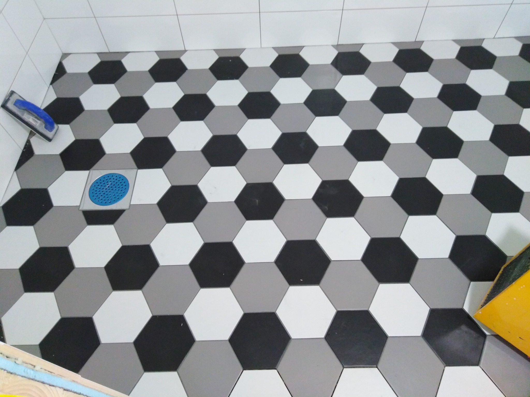 Ułożona podłoga w łazience z heksagonalnych płytek w odcieniach szarości, czerni i bieli, widoczna niebieska kratka odpływowa i narzędzia glazurnicze.