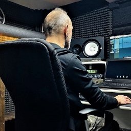 Mocny Głos Jerzy Janik - Mężczyzna w studio nagrań, siedzi przed komputerem z oprogramowaniem audio. Widoczne monitory studyjne, magnetofon szpulowy i panele akustyczne na ścianach.