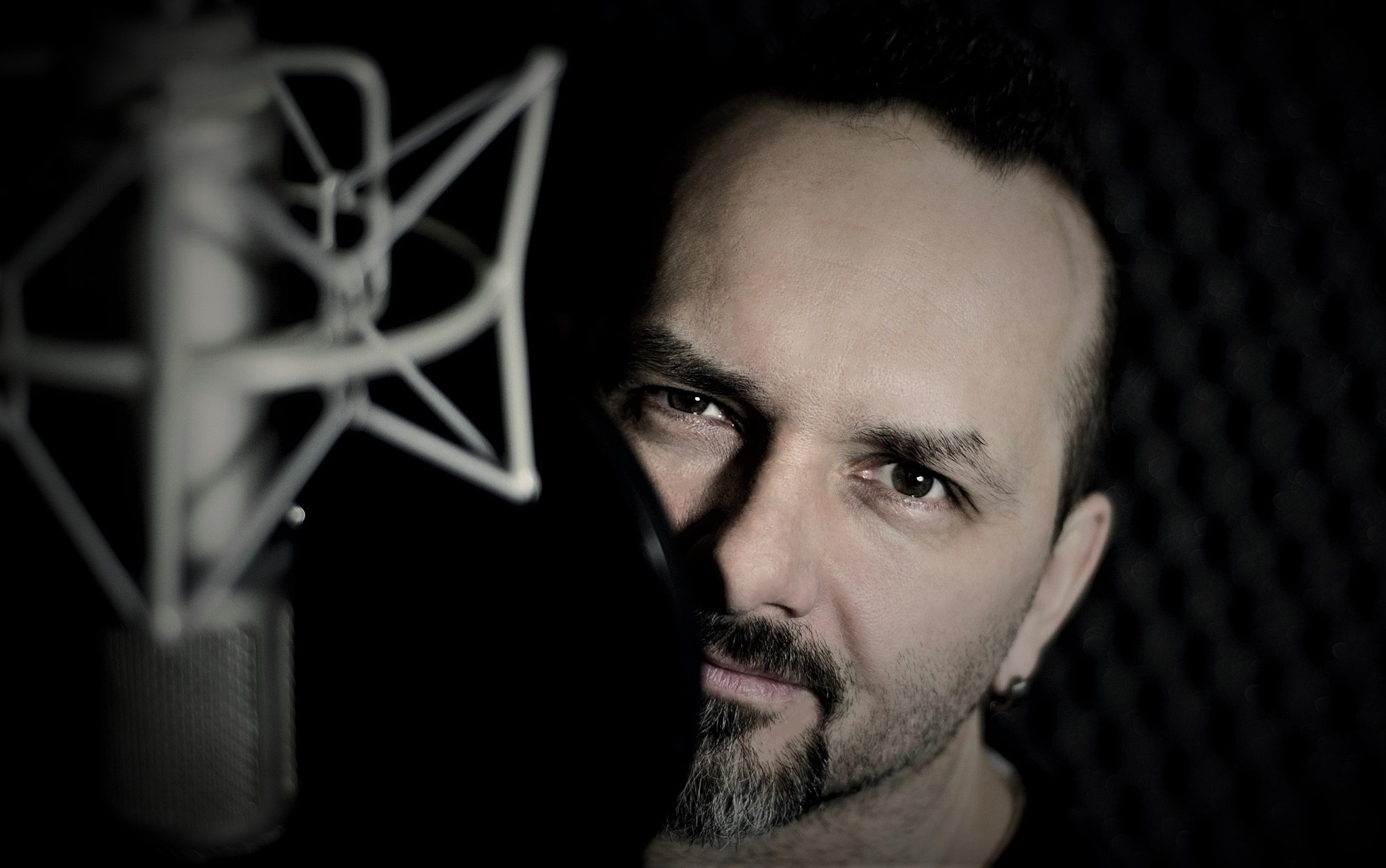 Portret mężczyzny w studiu nagrań w Krakowie, z mikrofonem studyjnym i wygłuszeniem akustycznym w tle. Ciemne oświetlenie, skupienie na twarzy lektora.