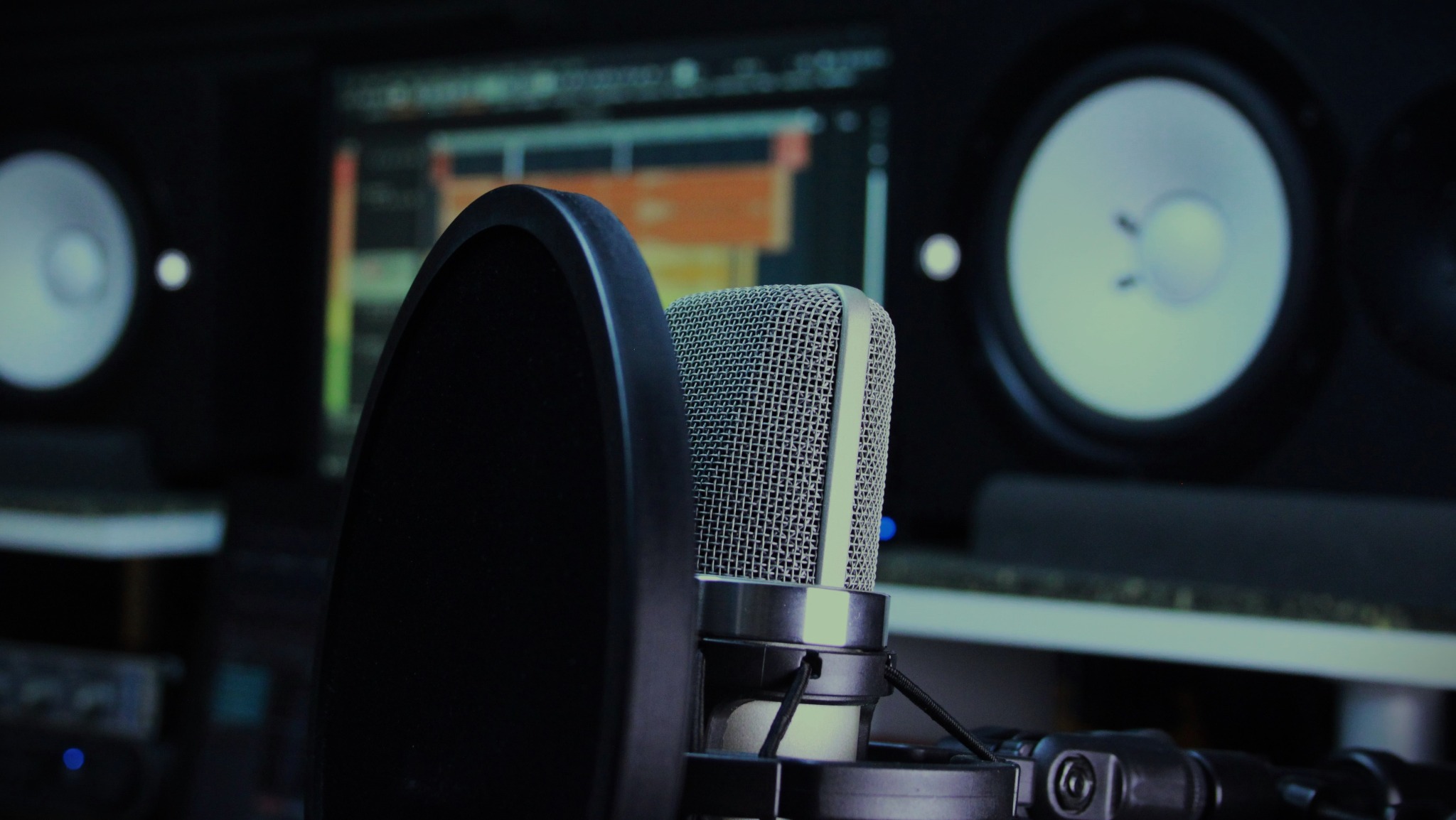 Mikrofon studyjny z pop filtrem na tle monitora z oprogramowaniem DAW i głośników studyjnych. Sprzęt w ciemnym pomieszczeniu studia nagraniowego.