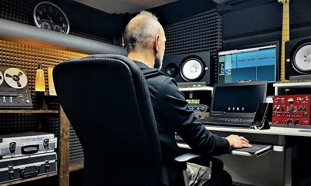 Mężczyzna w studio nagrań, siedzi przed komputerem z oprogramowaniem audio. Widoczne monitory studyjne, magnetofon szpulowy i panele akustyczne na ścianach.